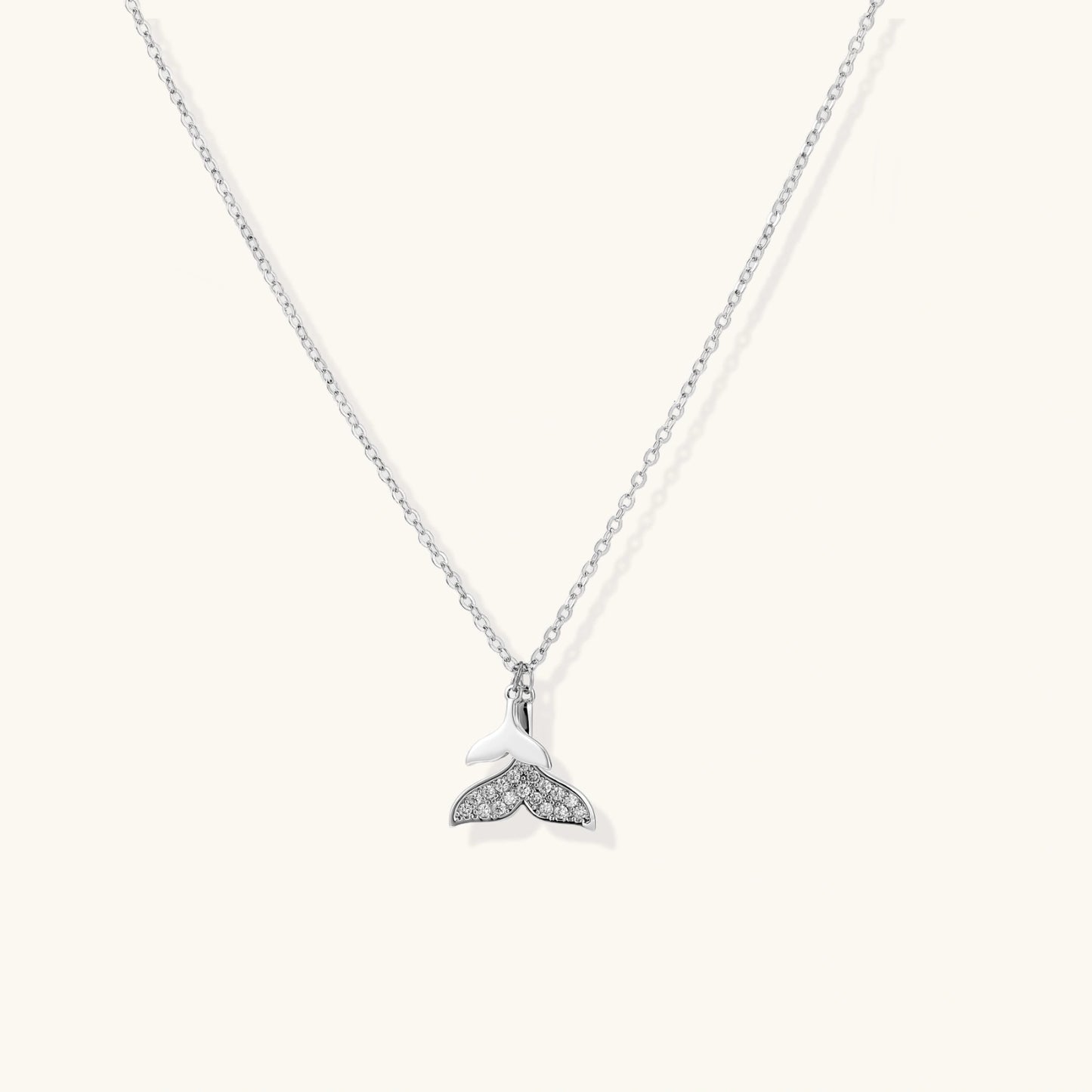 Delicate white gold plated whale tail pendant with pavé crystals on silver chain against cream background.