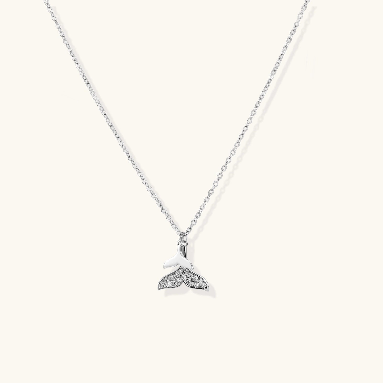 Delicate white gold plated whale tail pendant with pavé crystals on silver chain against cream background.